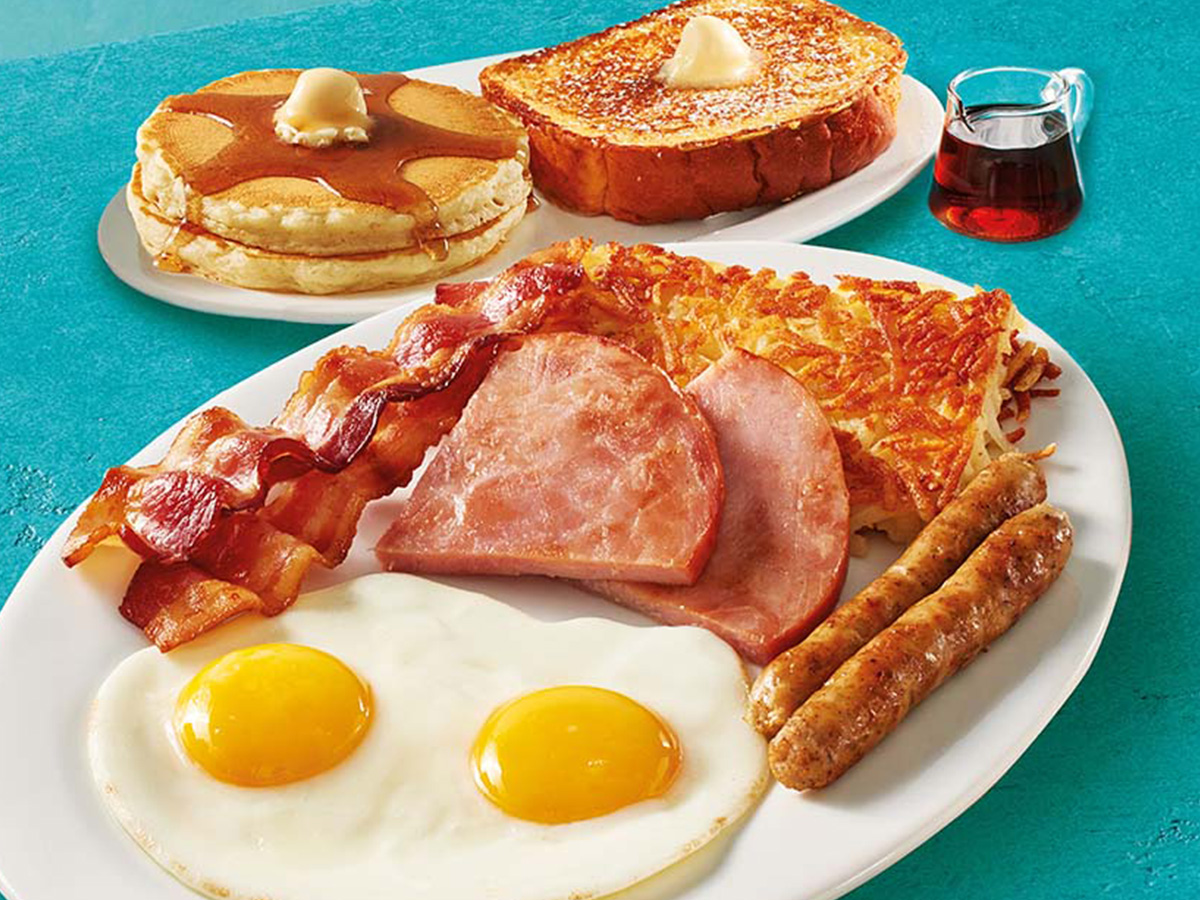 A plate with two eggs, hash browns, two slices of ham, two bacon strips, and two pork sausage links. A second plate has two buttermilk pancakes and a slice of Thick ‘N Fluffy French Toast.