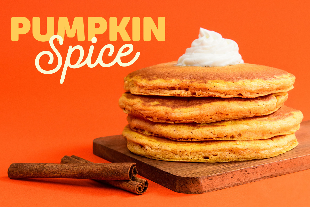 A full stack of Pumpkin Spice Pancakes with whipped topping on a wooden cutting board. Two cinnamon sticks are beside it.