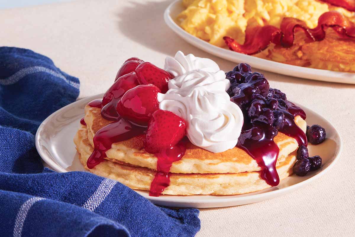 The Red, White, and Blueberry pancake combo, featuring two pancakes topped with strawberries, whipped topping, and blueberries, and a plate of scrambled eggs, bacon, and hash browns