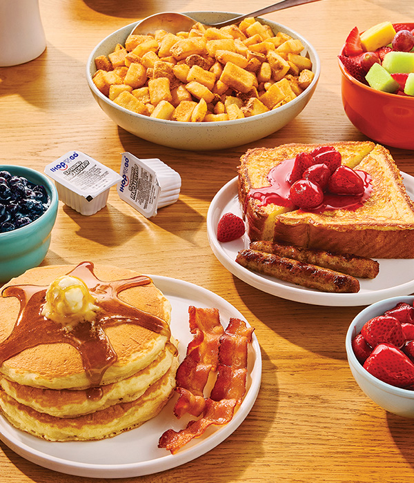 Plates of IHOP food, including buttermilk pancakes topped with butter and syrup, bacon, Thick 'N Fluffy French Toast, and crispy potatoes.