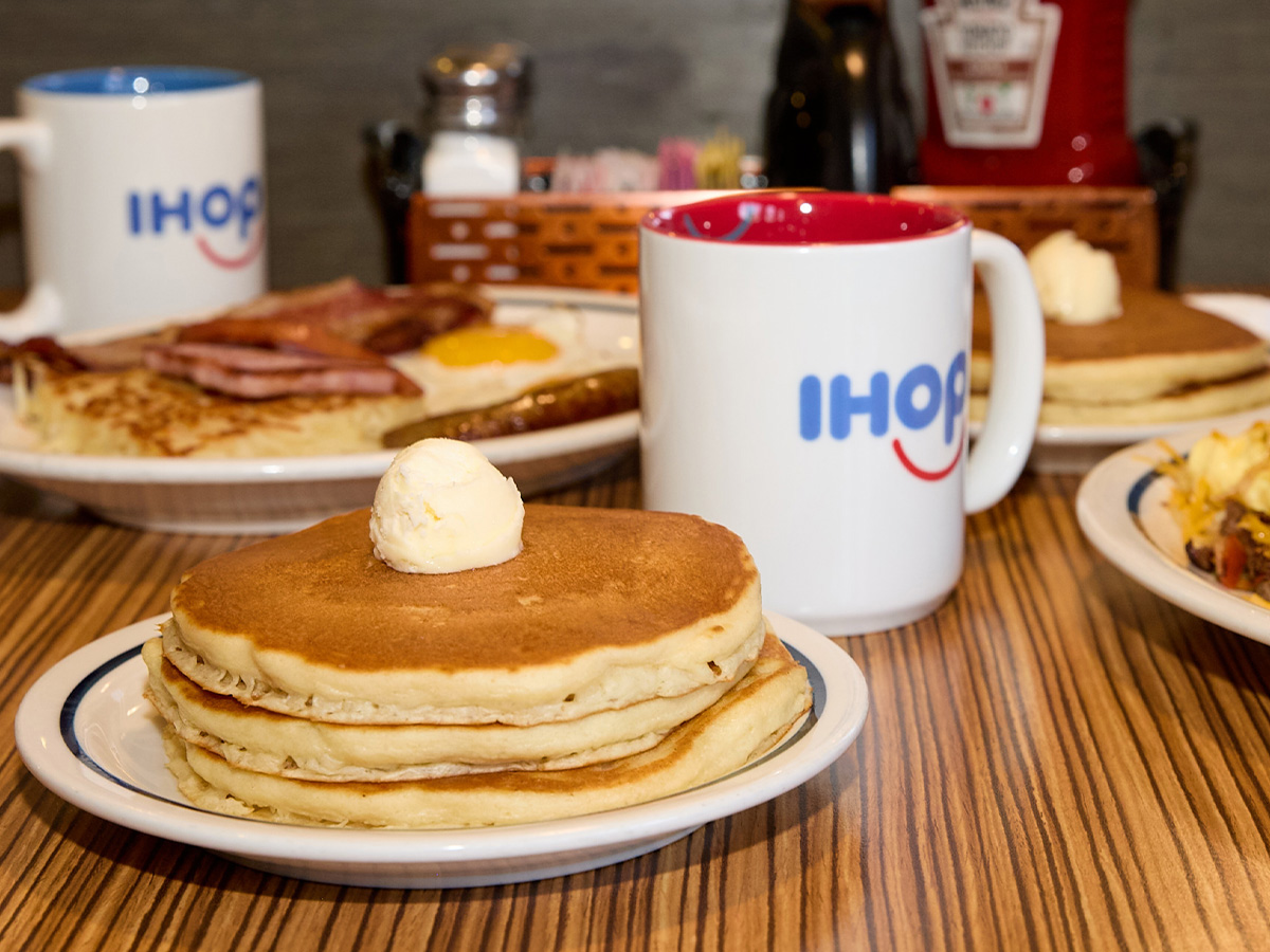 A stack of pancakes topped with butter on a plate, set on a table in a booth at IHOP. In the background is a mug of IHOP coffee and some breakfast meats.