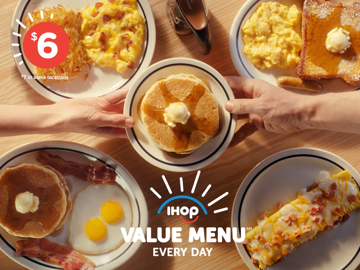 An assortment of IHOP Value Menu items on a wood table, with two hands reaching across and holding a plate of pancakes topped with syrup and buttter.