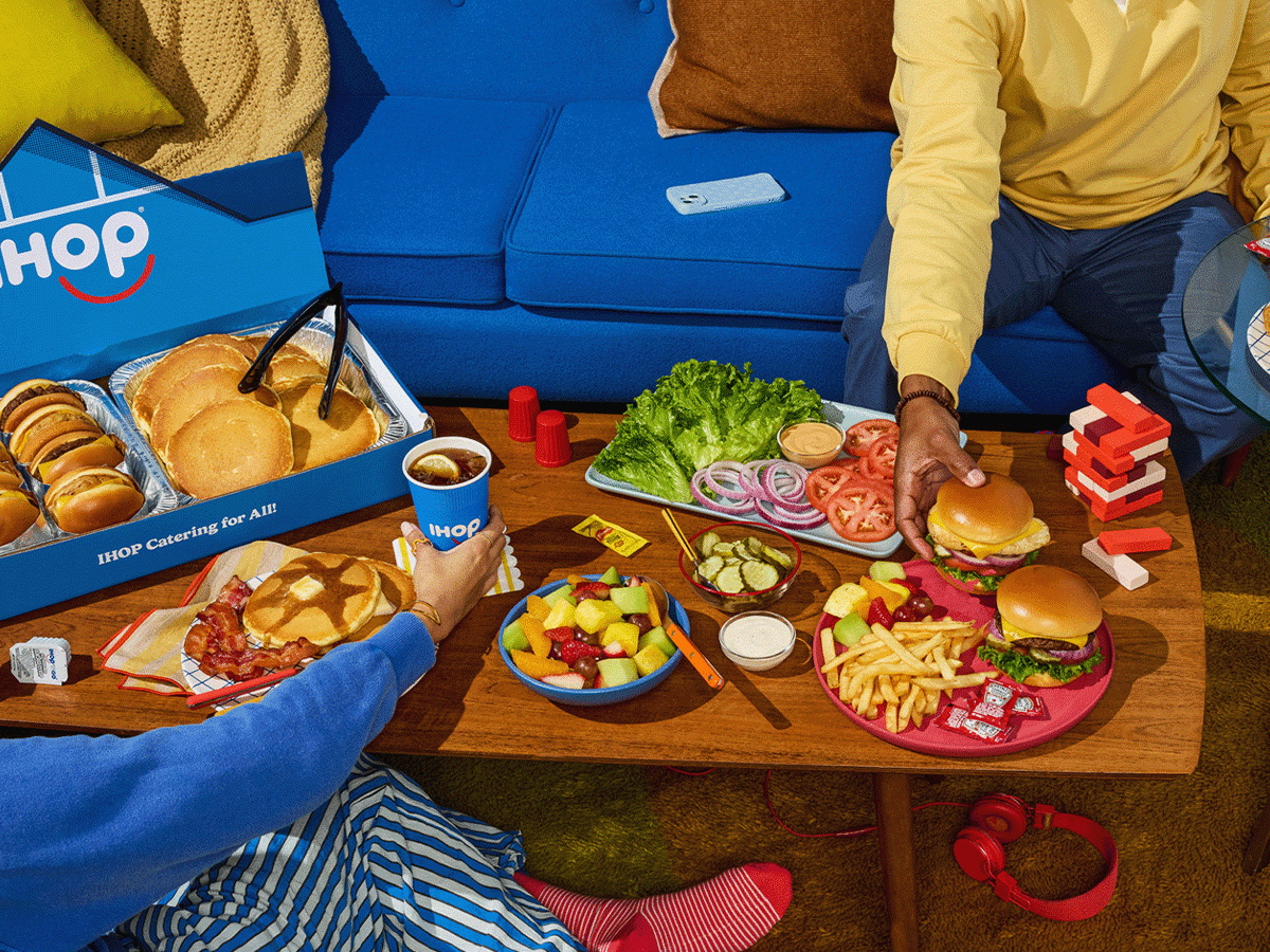 A scene in someone’s home, with a spread of IHOP Catering items laid out on a living room coffee table. Two people who are partially out frame grab items from the table.