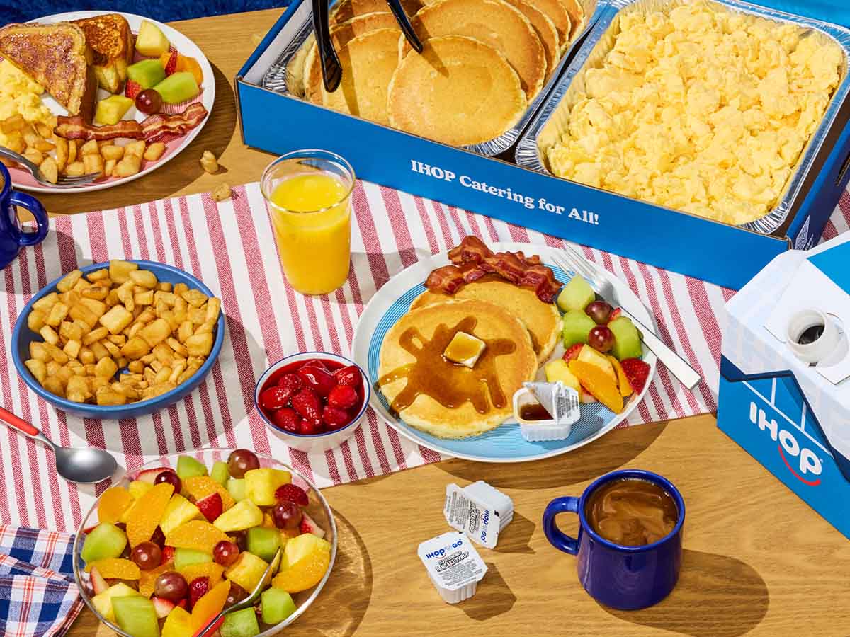 A spread of IHOP catering on a wood table with striped runner. Food includes a tray of pancakes, scrambled eggs, and mixed fruit.