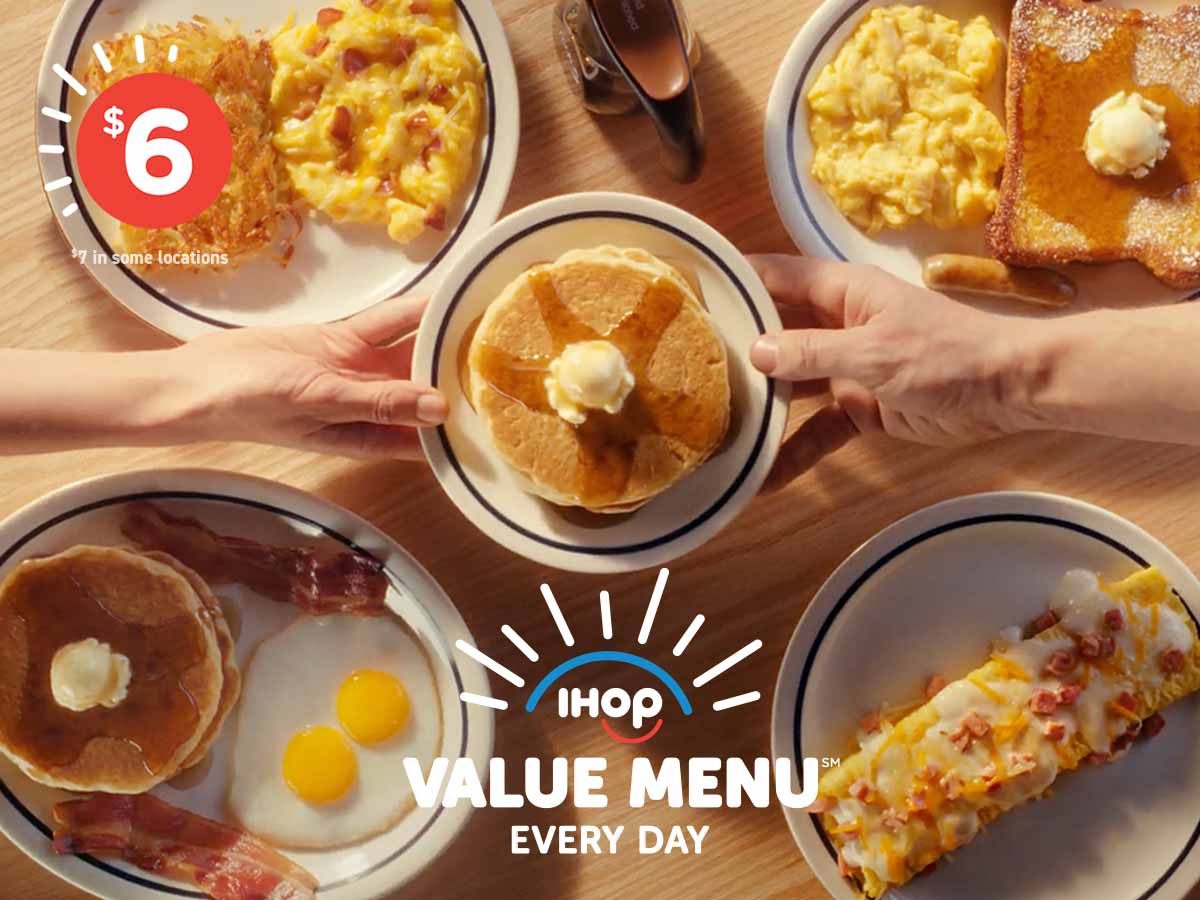 Two hands reach across a table, holding a plate of pancakes topped with butter and syrup. Beneath that on the table is a spread of IHOP Value Menu items.