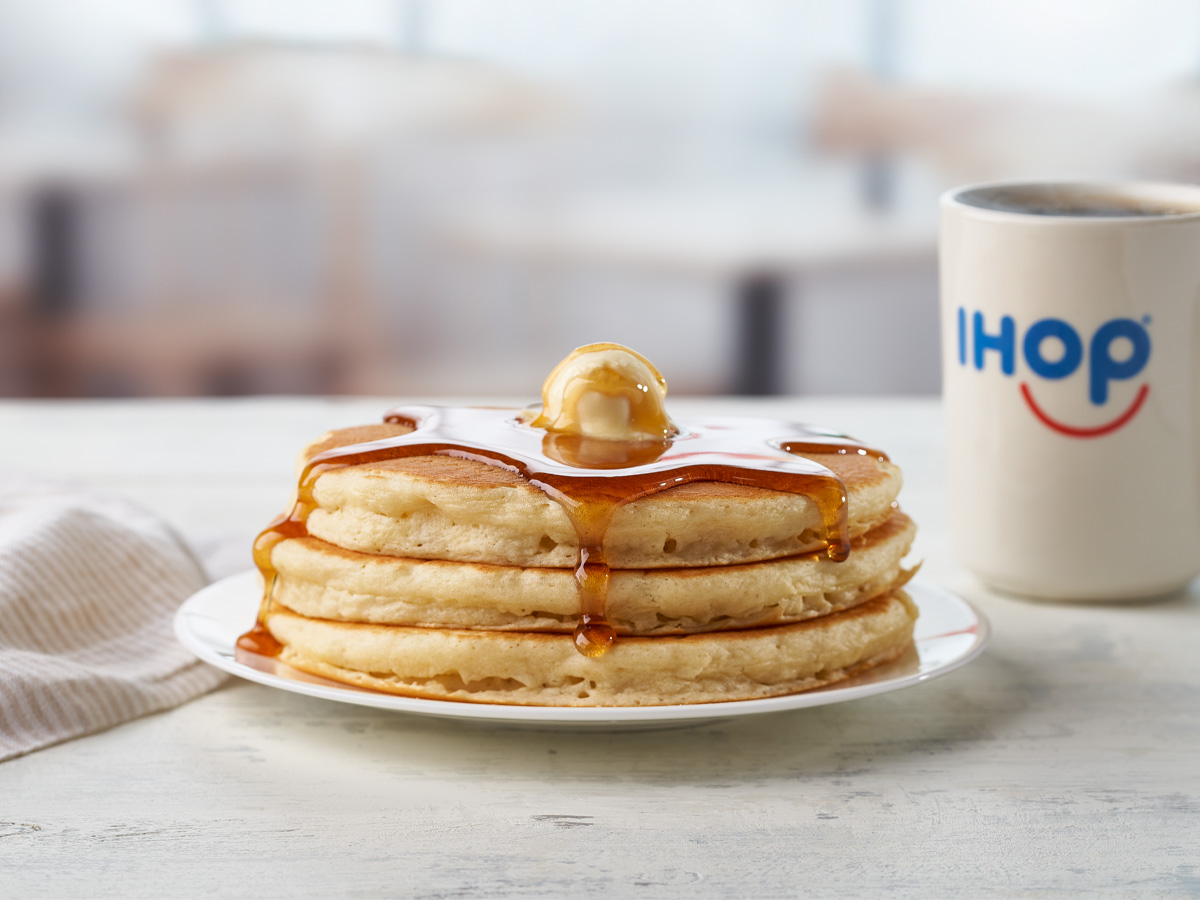 A short stack of IHOP pancakes on a plate, topped with butter and syrup. A mug of coffee is beside it.