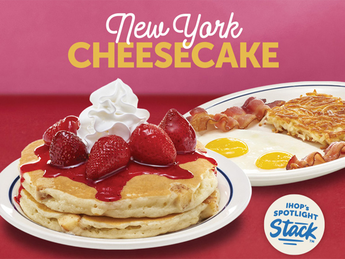 A plate of New York Cheesecake Pancakes with strawberries and whipped topping, alongside a plate of sunny side up eggs, two bacon strips and hash browns.