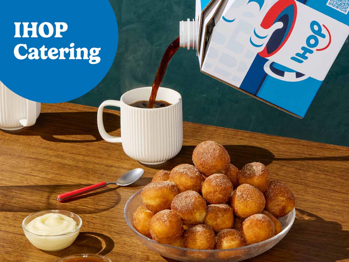 A catering box of IHOP coffee pours into a mug on a wooden table. A bowlful of Cinn-A-Dippers is also on the table beside a ramekin of dipping sauce.