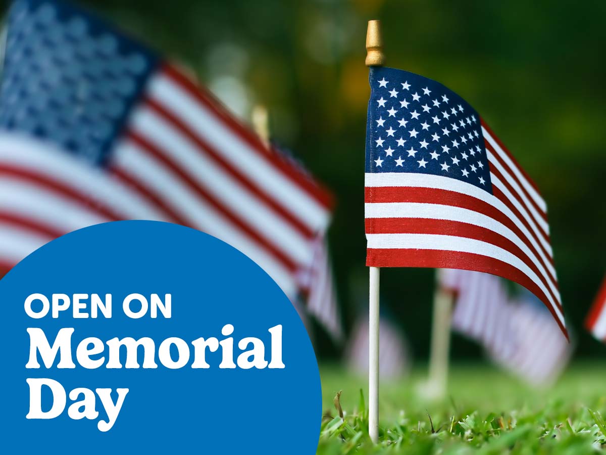 A small American flag planted in the grass, with inset text in a blue circle that says Open on Memorial Day