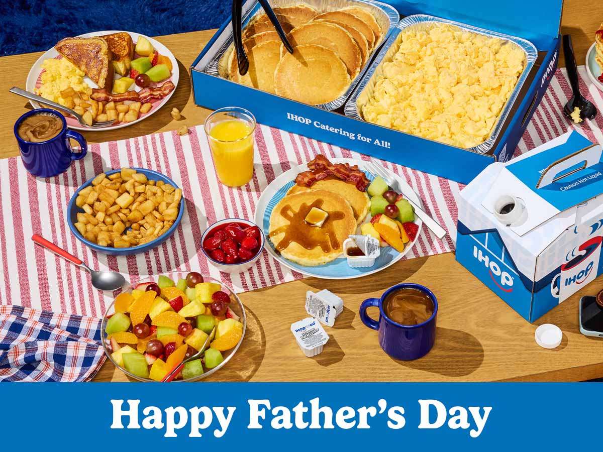 An arrray of IHOP catering on a wood table with a red and white striped runner. Food includes tins of pancakes and scrambled eggs, a box of coffee, and a bowl of fresh fruit