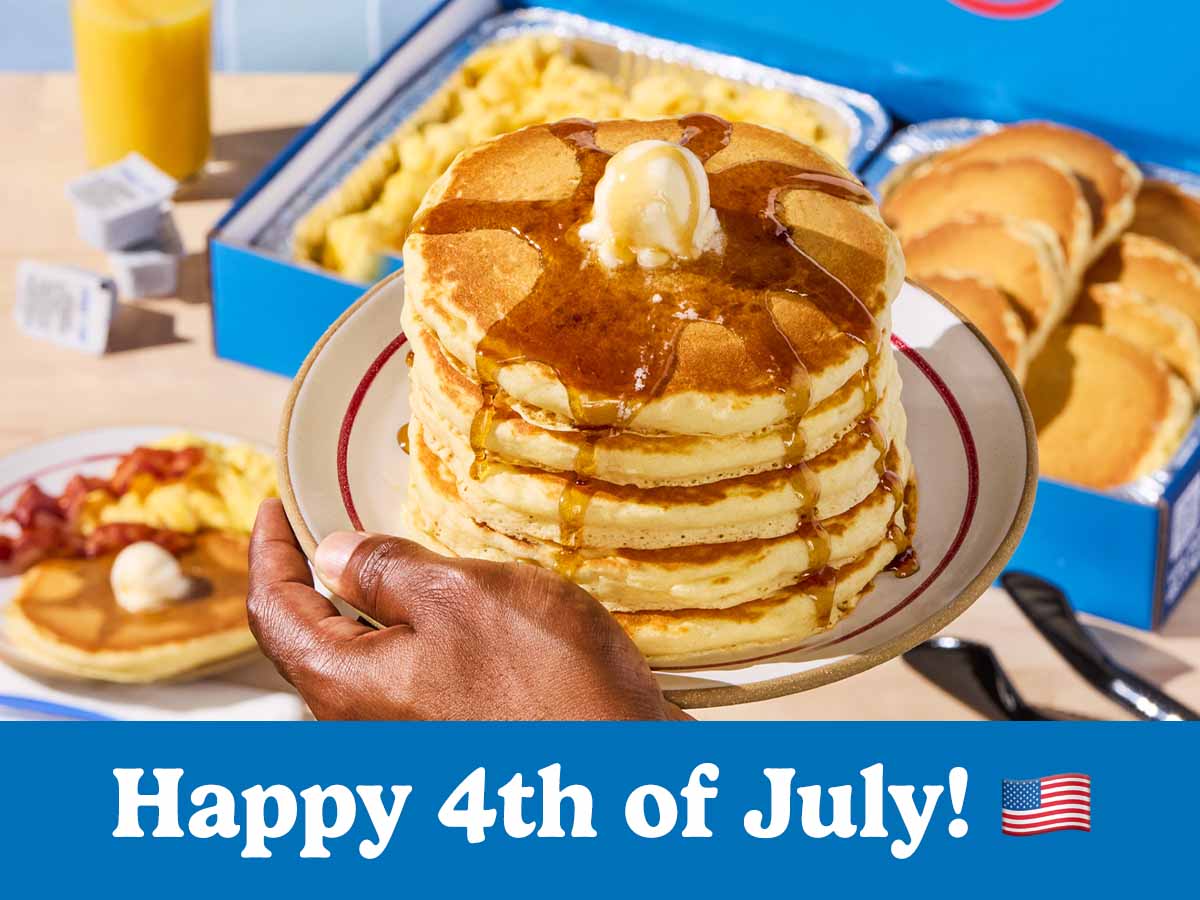 A stack of pancakes on a plate is being held above a catering tray of pancakes and scrambled eggs on a table below it