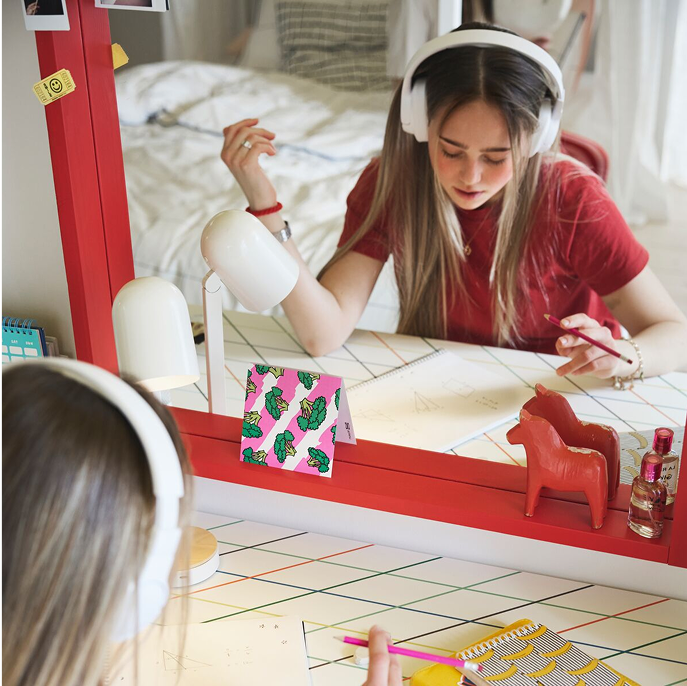 A teenager sitting studying by a LAGKAPTEN/SPÄND desk with a colorful grid pattern placed in front of a big red mirror.