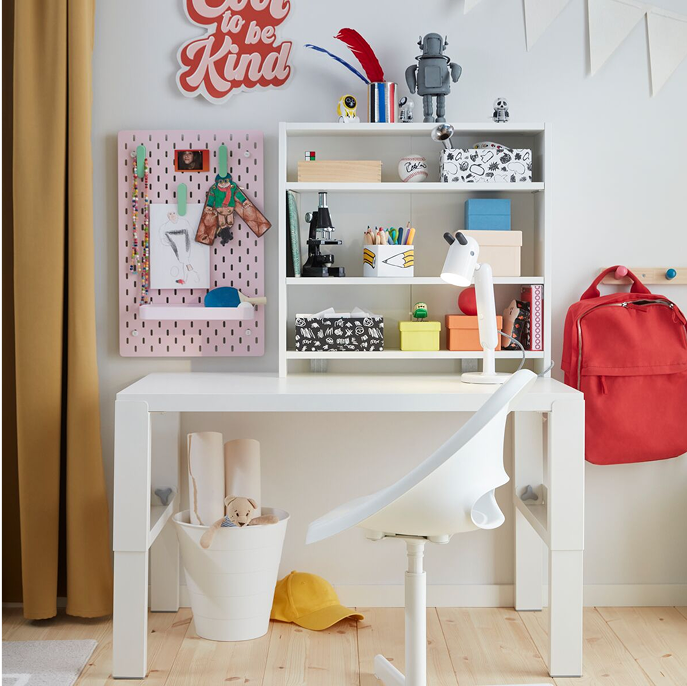 A white PÅHL desk and a white desk chair placed in a children’s room with a shelf and pegboard holding colorful objects. 