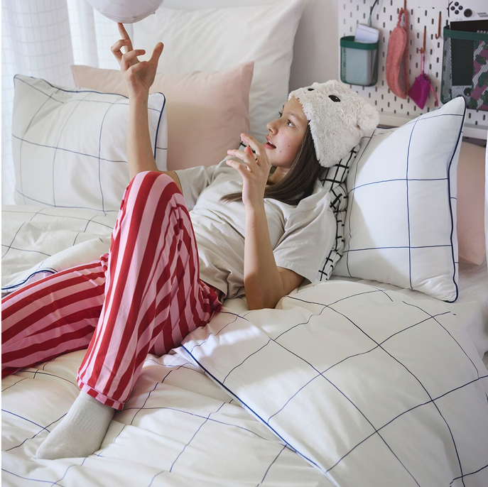 A teenager playing with a round object lying in a bed made with a BREDVECKLARE duvet cover in white with a blue grid pattern.