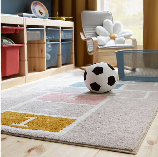 A STÄPPSPARV rug featuring a hopscotch pattern placed in a children’s room with a textile soccer ball and mesh storage units. 