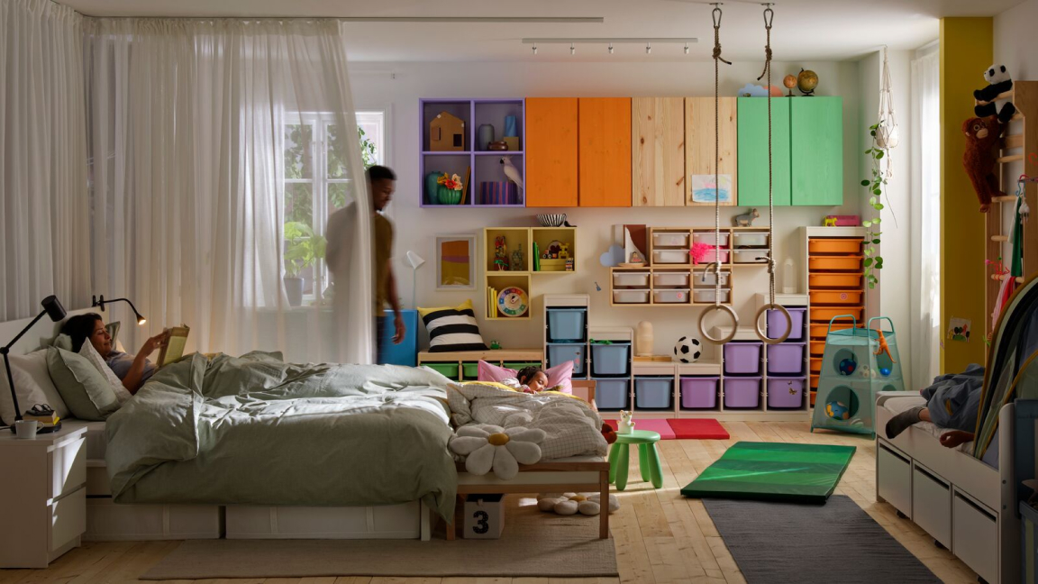 A room with two adults and a child. The child and one adult lie in separate beds. The other adult is standing by a curtain.