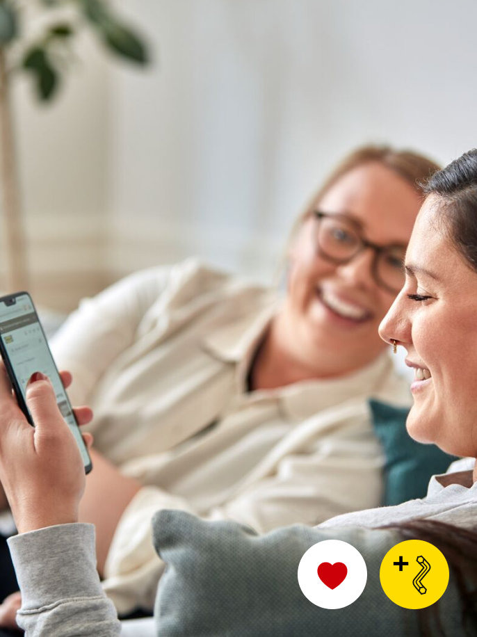 Two people smiling while one of them looks at the IKEA app on a phone. In the bottom-right corner, two icons – one of a red heart in a white circle, one of a black Allen key in a yellow circle.