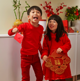 Two children wearing red outfits smile joyfully, holding festive decorations. The background features a cabinet with vases and red ornaments, suggesting a celebration.