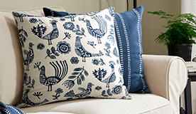 The KRUTBRÄNNARE cushion cover has an off-white background with a vibrant, rustic design of blue flowers, leaves and birds.