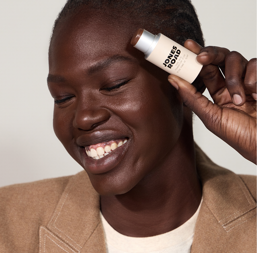 Close-up portrait of a smiling person holding a JONES ROAD beauty stick makeup product against their forehead