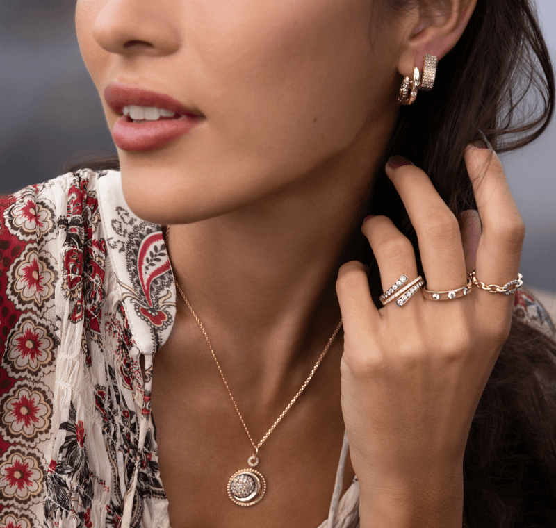 Woman wearing layered gold necklaces, gold hoop earrings, and stacked rings with a patterned blouse.