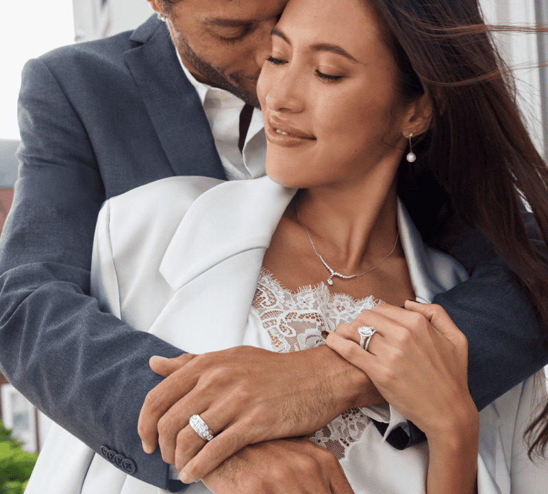 Couple embracing, woman wearing a diamond engagement ring and matching wedding band, shown close up on hands.
