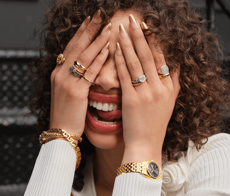 Model covering face playfully, wearing stacked gold rings, bracelets, and a gold watch.