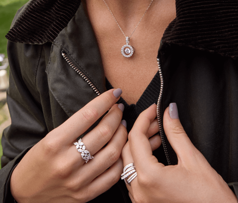 Model wearing a diamond halo pendant necklace and stacked diamond rings, shown close up against dark jacket.