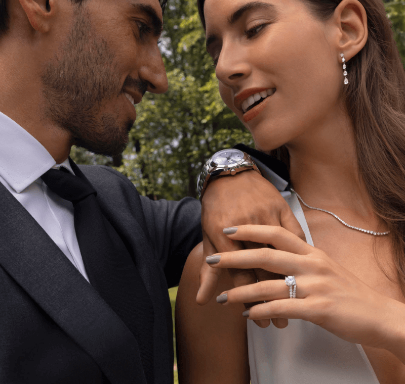 Smiling Couple Outdoors With Woman Showing Diamond Engagement Ring And Bridal Jewelry, While The Man Is Shown Wearing a Watch.