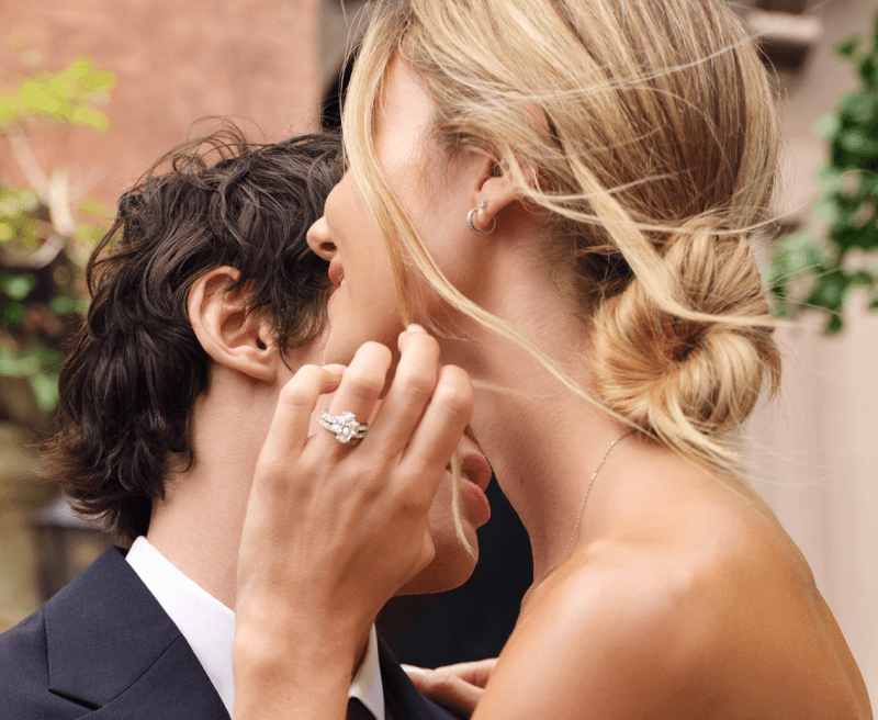Woman Wearing a White Gold Diamond Engagement Ring and Hoop Earrings Leaning Close to a Man in a Suit.