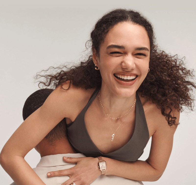 Woman on a man’s shoulders wearing yellow gold hoops, a chain necklace, a diamond drop pendant, and yellow gold-tone watch.