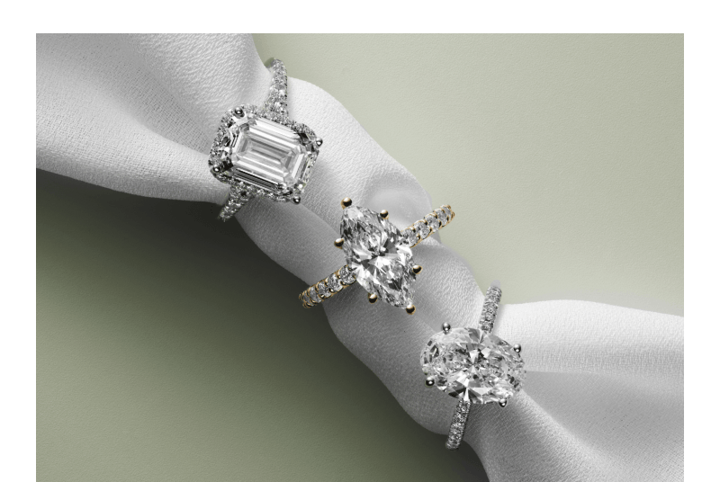 Three diamond rings featuring emerald, marquise, and oval cuts on pavé bands, styled on soft fabric.