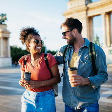 A smiling man and woman walk together outdoors on a sunny day, holding coffee cups.