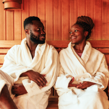 A man and woman in white bathrobes sit together in a wooden sauna, smiling at each other.