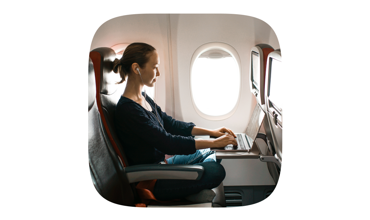 A woman sitting in an airplane window seat, working on a laptop placed on the tray table..