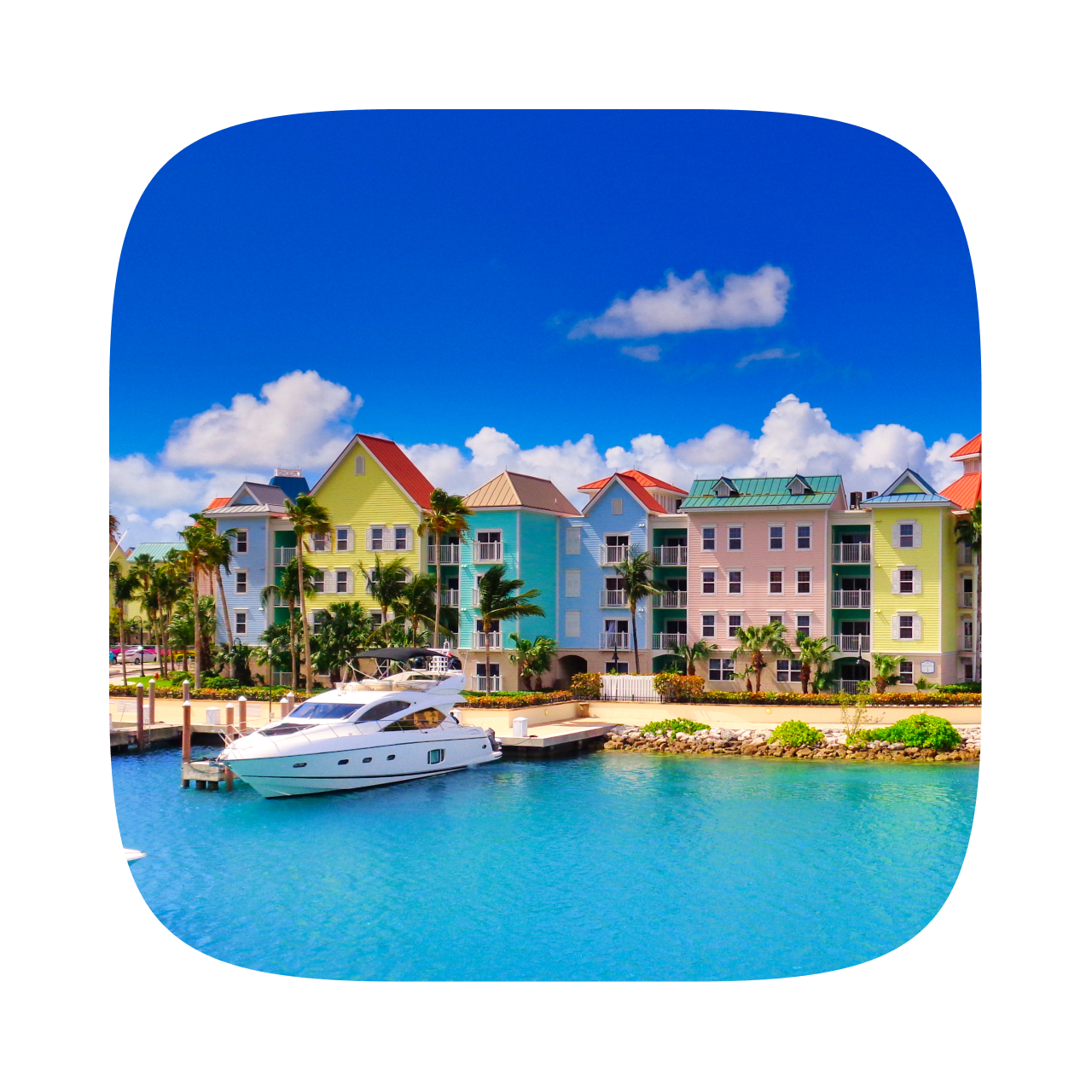 A white yacht is docked in a calm, turquoise harbor in front of a row of vibrant, pastel colored buildings.