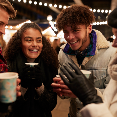 A group of young friends smiles and chats while holding warm drinks at an outdoor night market.