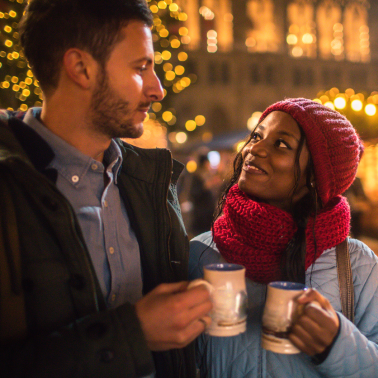 A man with a short beard and a woman wearing a red knit hat and scarf smile warmly at each other while holding mugs.