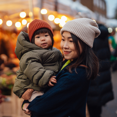 A woman wearing a light colored knit beanie smiles warmly as she holds a young child.