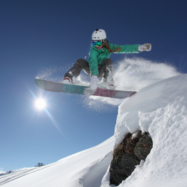 A snowboarder in a green jacket catching air off a snowy jump on a sunny day.