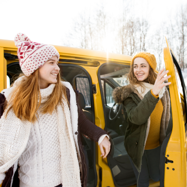 Two smiling women wearing winter hats and sweaters standing next to a bright yellow vehicle.