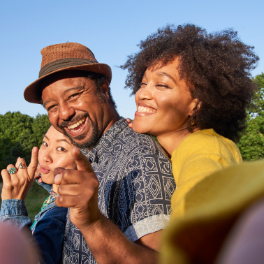 Three people, including a man in a fedora, are smiling happily in an outdoor setting with trees in the background.