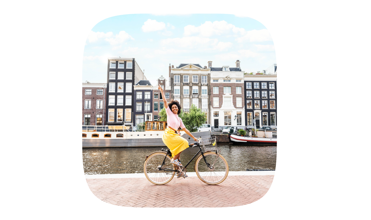Person riding a bicycle and waving beside a canal with historic canal houses behind.