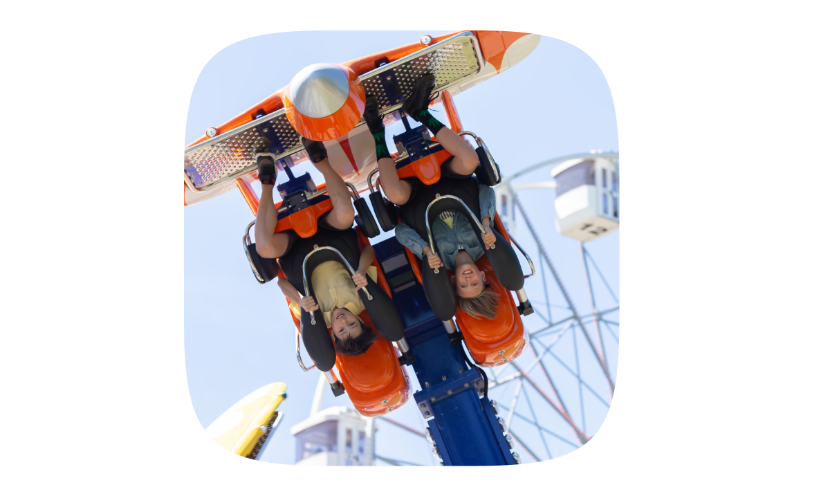 Two young boys hang upside down side-by-side high in the air at an amusement park, a Ferris wheel visible behind them.