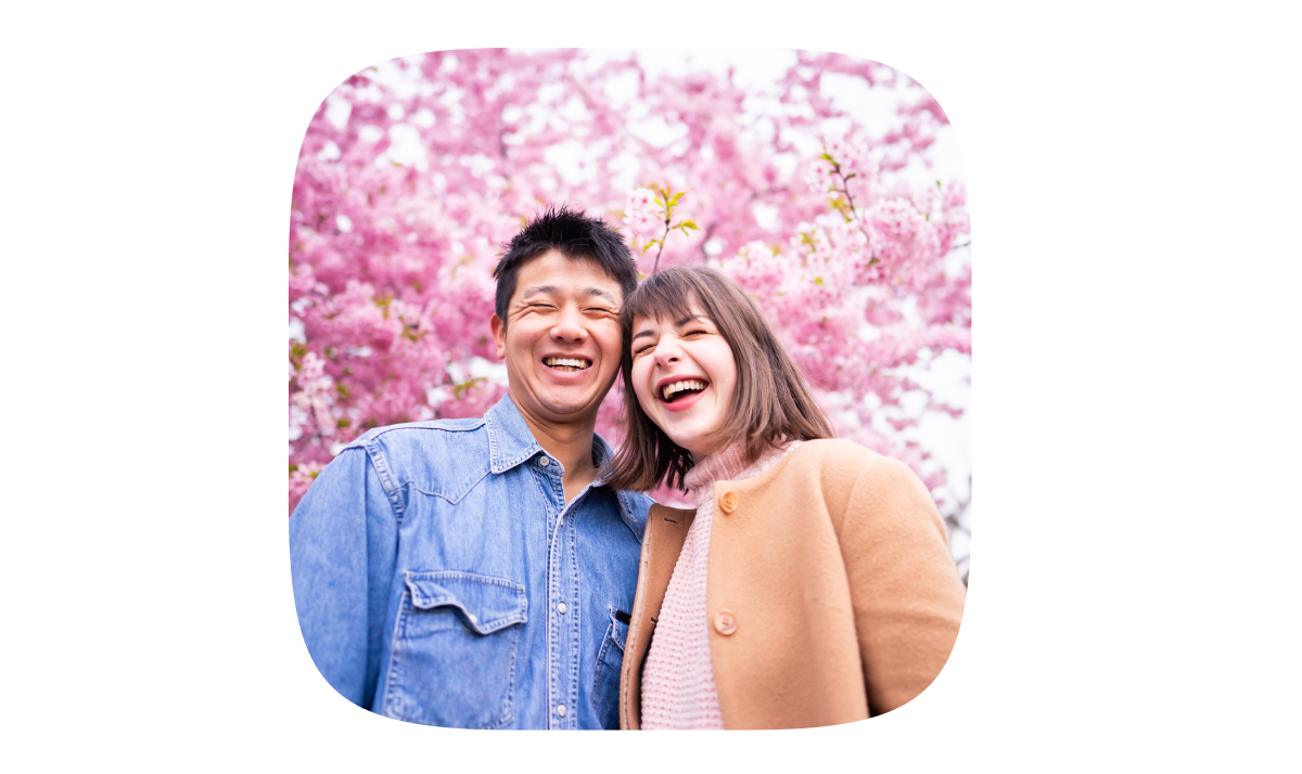 A couple is taking a photo, smiling, with cherry blossom trees in the background.