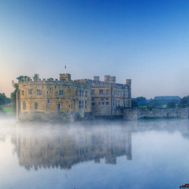 A large, historic stone castle surrounded by misty water.