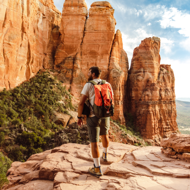 A man with a backpack stands on a rocky ledge overlooking red rock formations.