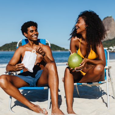 Couple smiling on beach chairs, man eating snacks, woman holding a coconut.