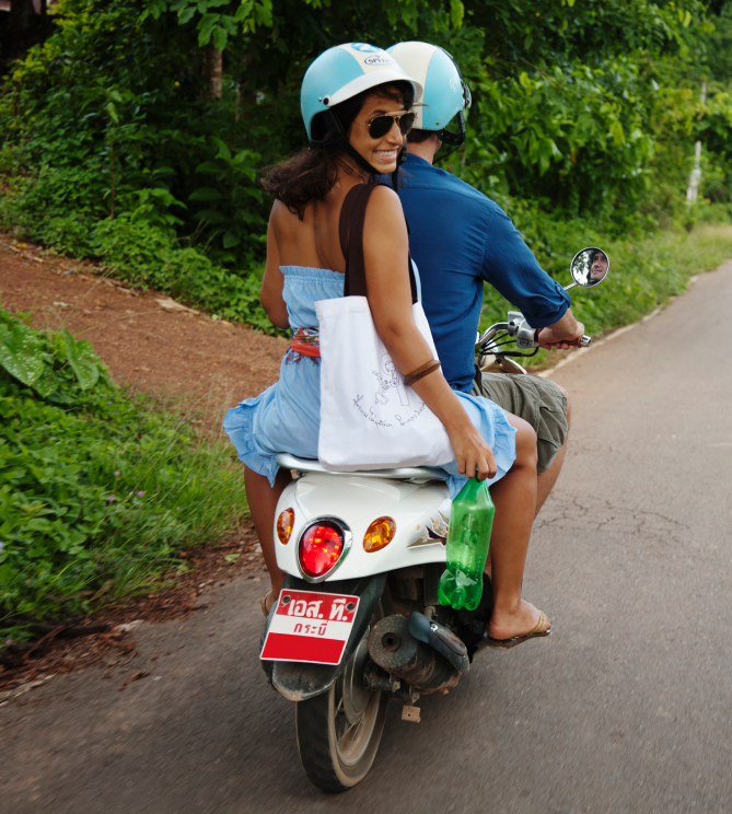 Two people riding a scooter on a rural road, the passenger looking back and smiling.