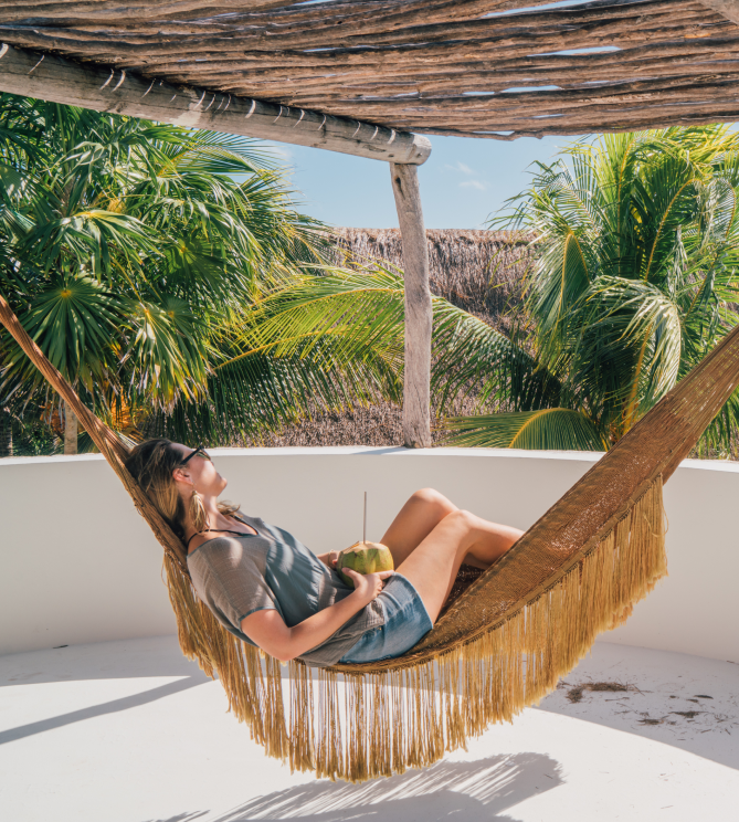 Person relaxing in a hammock under palms, holding a coconut drink.